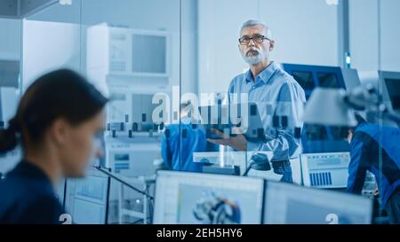 Modern Factory: Senior Engineer Looks at Camera, Holding Laptop, Inspecting, Overlooking Workshop Operation. Manufactory Has Professionals Working on Stock Photo