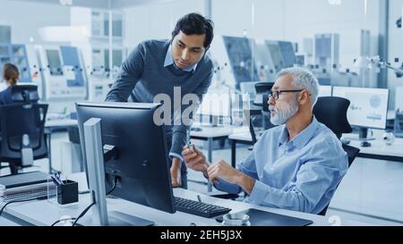 Modern Factory Office: Indian Project Manager Talks to Caucasian Senior Engineer whos Working on Computer, Inspect Metal Component. Professionals Stock Photo