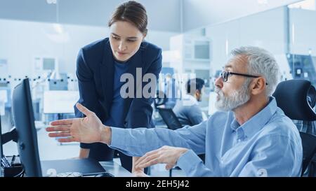 Modern Factory Office Young Confident Female Project Manager Talks to Senior Engineer Working on Computer. Team of Professionals Solving Heavy Stock Photo