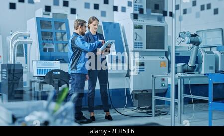 Factory Workshop: Professional Female Engineer and Male Machinery Operator Use Industrial Digital Tablet Computer to Program Robot Arm for Production Stock Photo