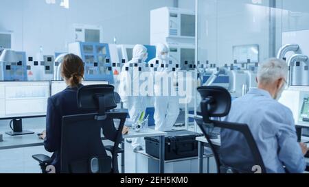 In Clean Workshop: Professional Workers in Coveralls and Masks Use Laptop and Talk. Inside Factory Office: Female Electronics Engineer Works on Stock Photo