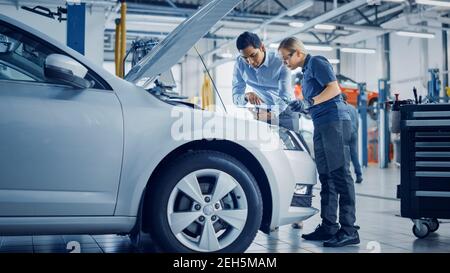 Manager Checks Tasts on a Tablet Computer and Explains an Engine Breakdown to an Female Mechanic. Car Service Employees Inspect Car's Engine Bay with Stock Photo