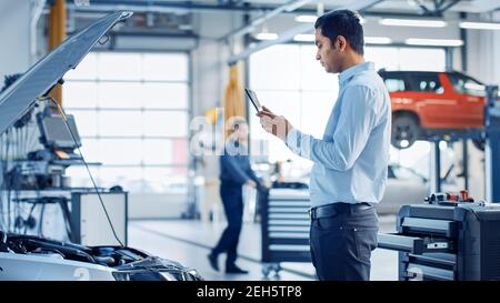 Car Service Manager Uses a Tablet Computer with an Augmented Reality Diagnostics Software. Specialist Inspecting the Car in Order to Find Broken Stock Photo