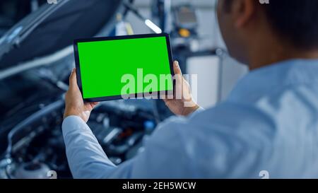 Car Service Manager or Mechanic Uses a Tablet Computer with a Green Screen Mock Up that is Pointed at an Enginer Bay. Specialist Inspecting the Stock Photo