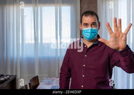 Bearded man in a purple shirt and surgical mask to protect himself from the coronavirus with a raised hand waving at the camera. Health concept Stock Photo