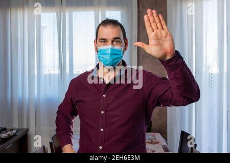 Bearded man in a purple shirt and surgical mask to protect himself from the coronavirus with a raised hand waving at the camera. Health concept Stock Photo