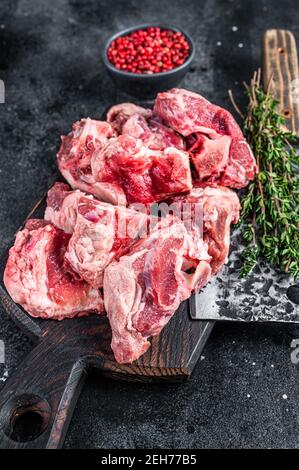 Raw lamb meat stew cuts with bone on wooden butcher board and cleaver. Black background. Top view Stock Photo
