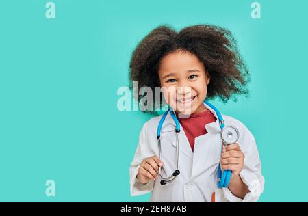 Portrait of positive African American female kid wearing a doctor coat with phonendoscope, standing on turquoise background Stock Photo