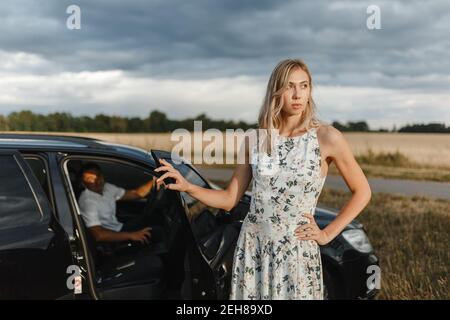 Blonde woman standing near the car door and man sitting inside the car. Couple in the car Stock Photo