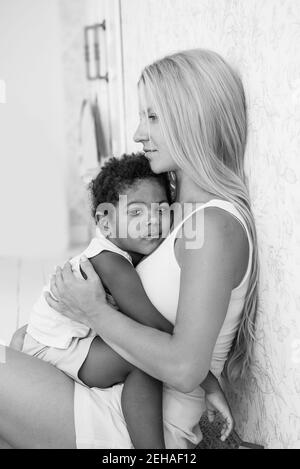 Portrait of a young blonde Caucasian mother holding, hugging little African American son in arms. The boy pressed against the womans chest. Tenderness Stock Photo