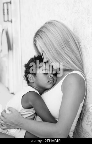 Portrait of a young blonde Caucasian mother holding, hugging little African American son in arms. The boy pressed against the womans chest. Tenderness Stock Photo