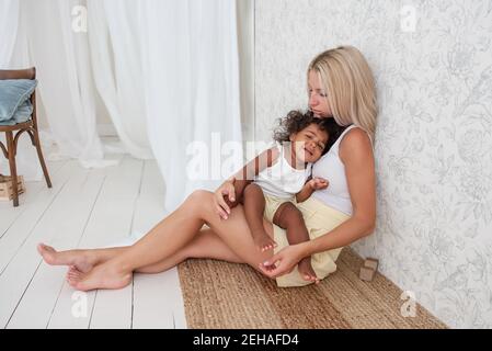 Portrait of young blonde Caucasian mother holding hugging little African American daughter in arms. Girl pressed against the womans chest. Tenderness Stock Photo