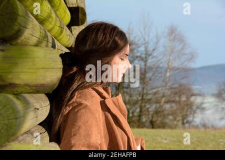 Young woman from the side, with light brown coat, leaning against a wooden hut, with a serious expression on her face and closed eyes, looks down,  en Stock Photo