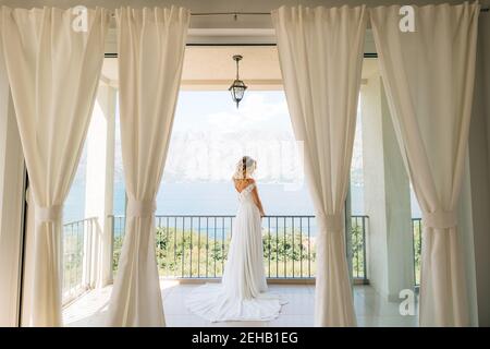 A sophisticated bride in an elegant wedding dress stands on a wide balcony with a picturesque view of the bay  Stock Photo