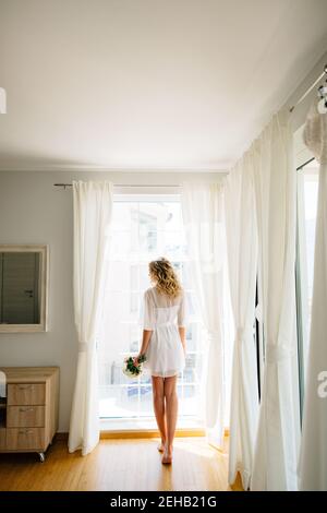 The barefoot bride in a white peignoir stands by the window with a wedding bouquet in her hands, back view  Stock Photo