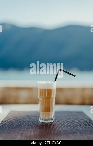 Faceted glass with coffee latte and a straw on a blue background, on a table in a cafe outside. A close-up of a creamy beverage with white foam. Stock Photo