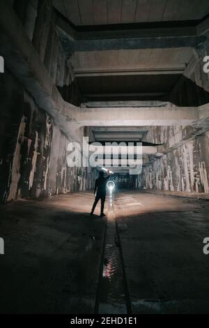 Back view of anonymous person with glowing flashlight in hand standing in abandoned tunnel with shabby walls Stock Photo