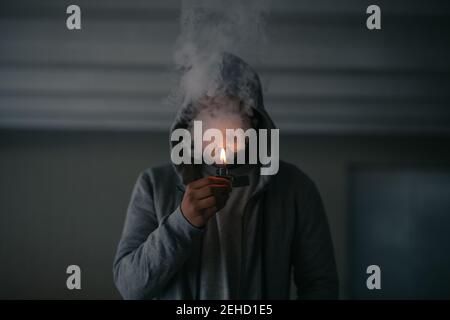 Unrecognizable male in hooded jacket smoking cigarette while standing in dark room with burning lighter in hand Stock Photo
