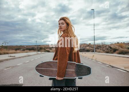 Side view of young female skater with long blond hair in trendy outfit standing on  asphalt road with cruiser skateboard in hand against cloudy sky in Stock Photo