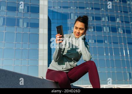 Happy young Hispanic lady in sportswear smiling and using mobile phone while resting on bench near modern building after outdoor training on sunny day Stock Photo