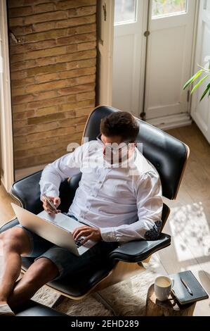 From above of male entrepreneur sitting in armchair and working online on project while using mobile phone and typing on laptop Stock Photo
