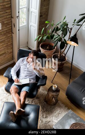 Businessman sitting in armchair and taking notes in organizer while talking on smartphone and checking messages Stock Photo
