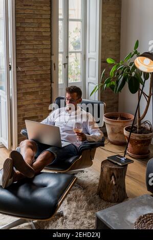 Handsome male entrepreneur sitting in armchair at home and working on project on laptop while enjoying wine Stock Photo
