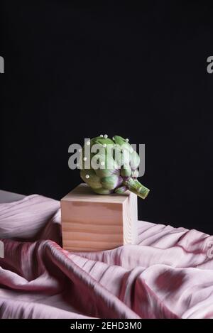 Ripe artichoke placed on wooden cube on table with silk fabric against black background Stock Photo