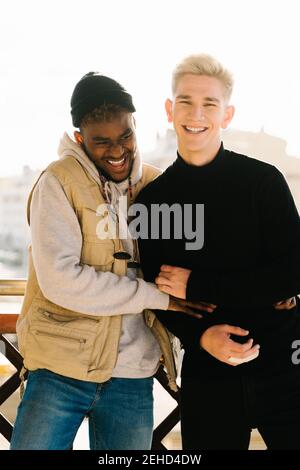 Joyful young multiracial gay couple in trendy outfits smiling and hugging while standing together on terrace on sunny day Stock Photo