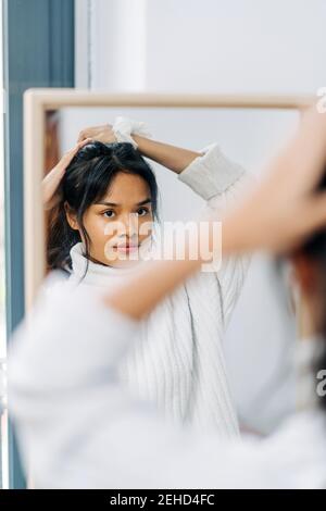 Long-haired brunette Asian Woman dressed in a white sweater, looking in the mirror and combing her hair Stock Photo