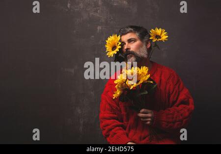 Creative mature male in knitted sweater covering face with bright sunflowers against black background Stock Photo