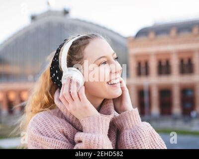 Side view of cheerful teenage female with long wavy hair in warm sweater smiling while enjoying music in headphones standing on city square Stock Photo