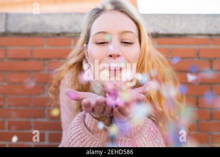 Happy young female with long blond hair blowing colorful confetti from hands while standing near brick wall on sunny day Stock Photo