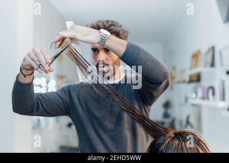 Concentrated young ethnic bearded male master cutting hair of crop unrecognizable customer in modern salon Stock Photo