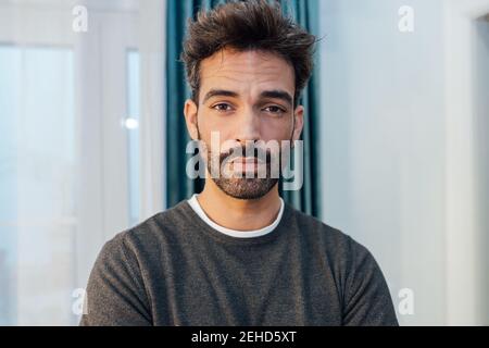 Concentrated young bearded ethnic male in casual clothes standing in modern apartment and looking at camera Stock Photo