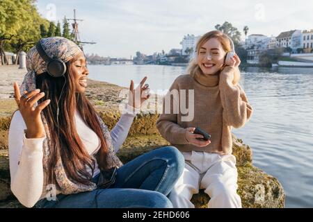 Positive multiracial female best friends sitting on embankment and enjoying songs in headphones with closed eyes while having fun together Stock Photo