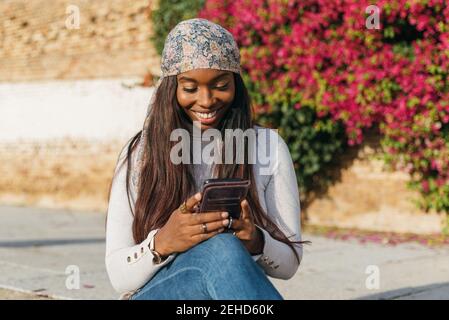 Confident smiling millennial African American female in stylish casual wear taking selfie on mobile phone while standing against wall of modern buildi Stock Photo