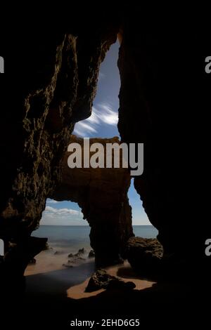 Through cave scenery view of rough rocky formations on sandy Pinhao Beach against sea under cloudy sky in Algarve Portugal Stock Photo