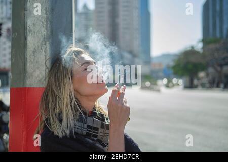 Side view of young female in scarf smoking cigarette near post on urban roadway in back lit Stock Photo