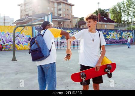 Two teenage boys with skateboard and backpack shaking hands and laughing on the street Stock Photo