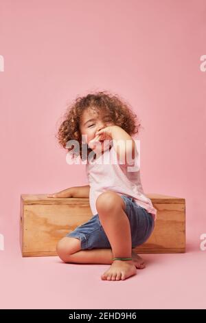 Charming cheerful barefoot child in t shirt and denim shorts with curly hair sitting on the floor playing on wooden platform Stock Photo