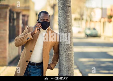 Unrecognizable thoughtful young black man in trendy outfit and face mask leaning on tree on city street and looking away while having phone conversati Stock Photo