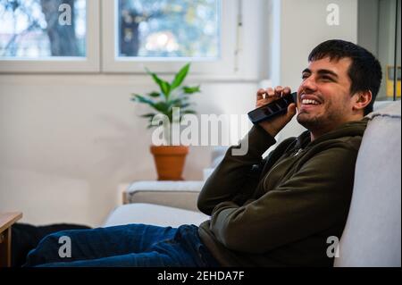 Side view of young smiling male suffering from blindness sitting on couch and listening audio message on mobile phone Stock Photo