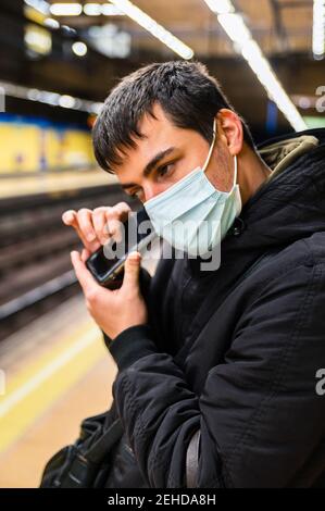 Side view of blind male passenger in medical mask waiting for train at platform and listening speaker on mobile phone Stock Photo