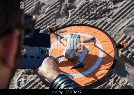 From above of unrecognizable male with remote controller standing on beach with drone placed on landing pad Stock Photo