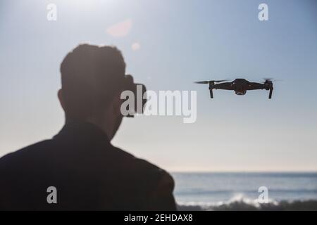 Back view of unrecognizable male wearing modern VR goggles operating drone with remote controller and experiencing virtual reality while standing agai Stock Photo