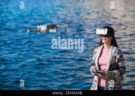 Excited female wearing modern VR goggles operating drone with remote controller and experiencing virtual reality while standing against blurred sea Stock Photo
