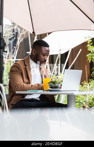 Young African American businessman browsing on laptop while looking down at cafeteria table Stock Photo