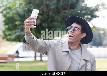 Cheerful ethnic stylish male wearing trendy outerwear and hat smiling widely and taking self portrait on cellphone standing near modern building Stock Photo