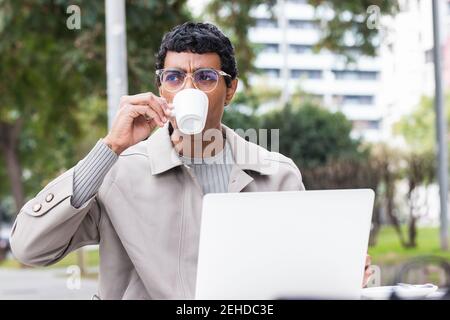 Concentrated African American male entrepreneur in stylish outfit and eyeglasses sitting in street cafe and browsing netbook while drinking beverage Stock Photo